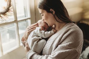 A young mother tenderly kisses her sleeping newborn baby while standing near a sunlit window.