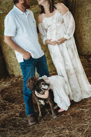 Couple in maternity photoshoot with dog, woman in floral dress holding baby bump, man in white polo and jeans outdoors.