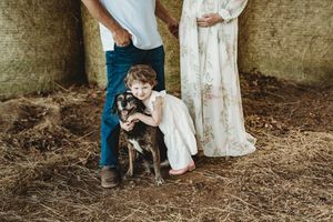 A young girl in a white dress hugs a dog beside adults in a rustic barn setting with hay on the ground.
