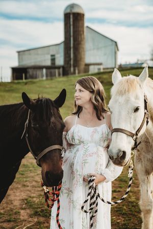 Pregnant woman in floral maxi dress standing between black and white horses on a farm with a barn backdrop.