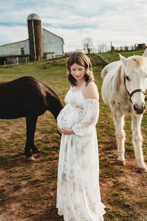 Pregnant woman in white floral dress cradling her belly beside black and white horses on a rustic farm.