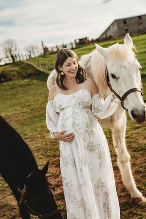 Pregnant woman in floral dress smiling beside a white horse on a green farm field.