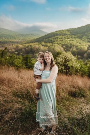 A smiling woman in a mint dress holds a toddler boy in a field with green mountains under a blue sky.