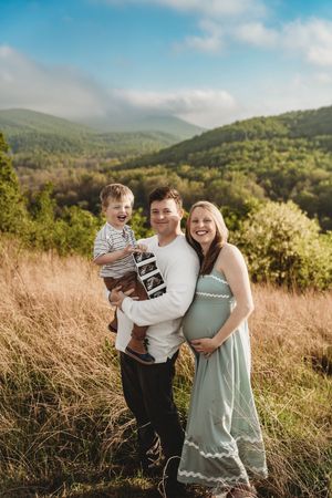 Smiling pregnant couple holding toddler son with ultrasound photos in scenic mountain meadow.