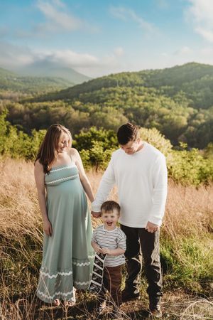 Pregnant woman in sage dress and husband hold hands with laughing toddler in a scenic mountain field.