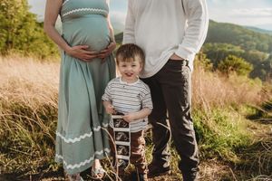 Pregnant woman in teal dress with toddler son and partner posing outdoors in grassy field with scenic hills.