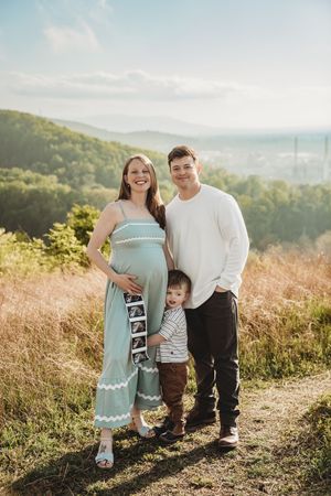 Pregnant woman in teal jumpsuit poses with husband and toddler son on scenic hilltop overlook during golden hour.