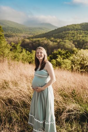 Smiling pregnant woman in a sage green maternity dress cradles her belly in a sunlit mountain meadow.
