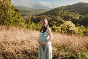 Pregnant woman in sage dress cradles baby bump in golden field with lush green mountains in background.