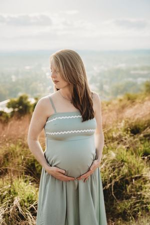 Pregnant woman in sage green dress cradling baby bump outdoors on a hillside with scenic landscape background.