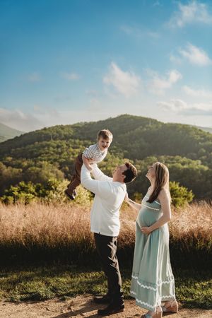 Happy couple lifting toddler in air during outdoor maternity photo shoot in mountain meadow.