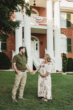 Pregnant couple holding hands in front of a brick colonial mansion with white columns and patriotic bunting.