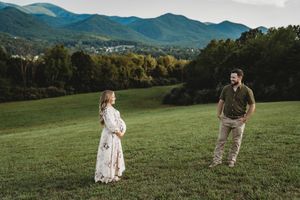 Pregnant couple standing apart in lush green field with stunning Blue Ridge Mountain backdrop during golden hour maternity session.