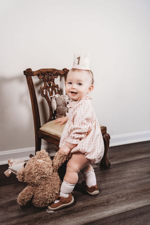 Smiling baby girl in pink romper and number 1 crown stands by vintage chair with teddy bear on first birthday.