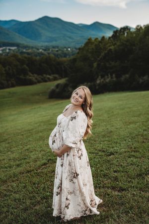 Smiling pregnant woman in floral maxi dress holding baby bump in green mountain meadow.