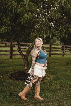 Pregnant woman with tattoos holding ultrasound photos, standing by a peach tree on a farm.