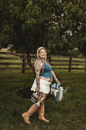 Tattooed pregnant woman holding watering can with flowers and ultrasound photo, standing in green field near wooden fence.