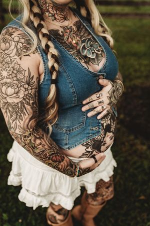 Heavily tattooed pregnant woman in denim dress cradling baby bump outdoors, showcasing full sleeve and body tattoos.