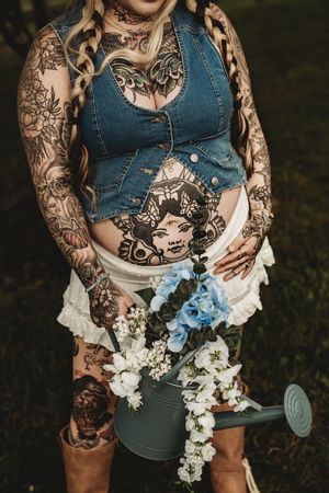 Heavily tattooed pregnant woman holding a watering can with blue and white flowers, wearing a denim vest outdoors.