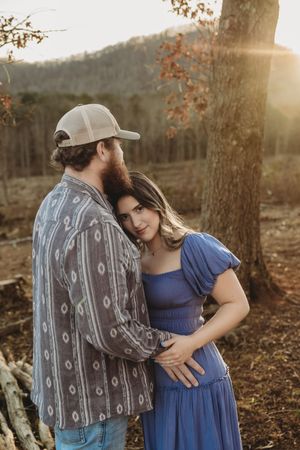 A couple embraces at golden hour near a tree, woman in blue dress, man in patterned shirt and cap, mountains behind.