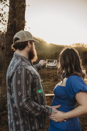 Couple sharing a pregnancy ultrasound photo outdoors at golden hour, woman in blue dress with baby bump.