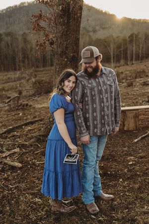 Couple holding ultrasound photo at golden hour outdoors, woman in blue dress, man in patterned shirt and cap.