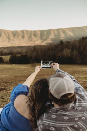 Couple lying outdoors holding ultrasound photo with mountain landscape backdrop, pregnancy announcement in nature.