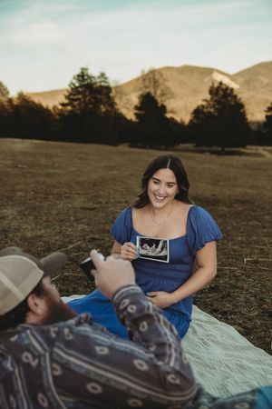 Happy pregnant woman in blue dress holds ultrasound photo while sitting on blanket in scenic mountain meadow.