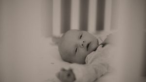 Black and white photo of a peaceful newborn baby lying awake in a white crib, looking upward softly.