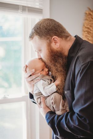 Bearded father tenderly kissing his newborn baby while standing near a bright window indoors.
