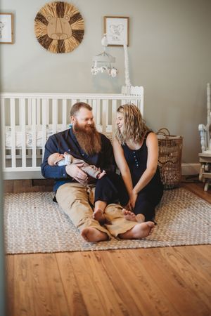 New parents sit on nursery floor with newborn, dad holding baby while mom smiles, white crib and lion decor behind them.