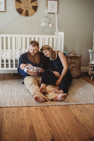 Happy couple sitting on rug in baby nursery, father cradling newborn while mother leans close, white crib in background.