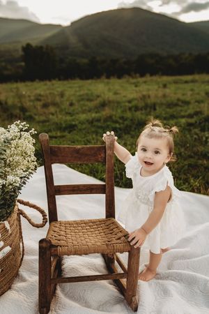 Smiling toddler girl in white dress standing by rustic wooden chair on white blanket with mountain meadow backdrop.