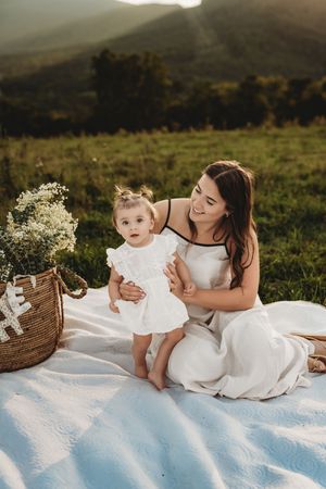 Mother and baby girl in matching white dresses sit on a blanket in a scenic mountain meadow at golden hour.