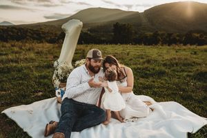 Family of three sitting on a white blanket in a green meadow with mountains at sunset, celebrating baby's first birthday.