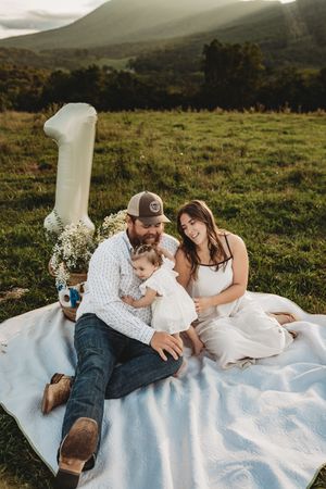 Family celebrating baby's first birthday on a blanket in a scenic mountain meadow with a number one balloon.