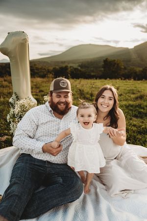 Happy family of three celebrate baby's first birthday outdoors in a mountain meadow at sunset.