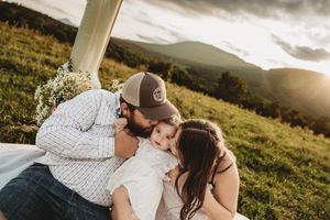 A couple kisses their baby on a grassy hillside at sunset, with mountains in the background.