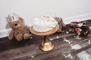 White frosted smash cake on gold pedestal stand with teddy bears and cake crumbs scattered on hardwood floor.