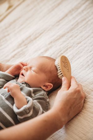 Parent gently brushing newborn baby's hair with a Hudson wooden bristle brush while infant lies on soft white blanket.