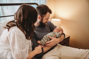 New parents sitting on bed admiring their newborn baby in a warmly lit bedroom.
