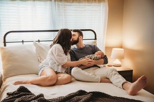 Young couple sitting on bed sharing a tender kiss while holding their newborn baby in a cozy bedroom.