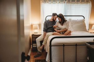 Couple sitting together on a bed near a window, sharing a tender moment in a cozy bedroom with warm lighting.