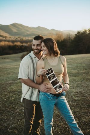 Happy couple holding ultrasound photos outdoors with mountain backdrop during golden hour pregnancy announcement.