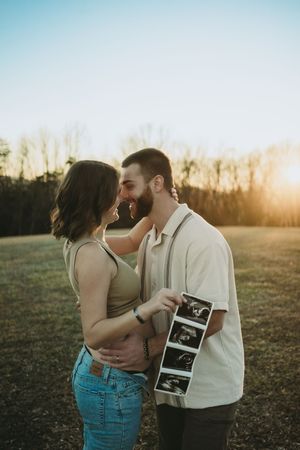 Couple sharing a tender moment at sunset in a field, man holding ultrasound photos announcing pregnancy.