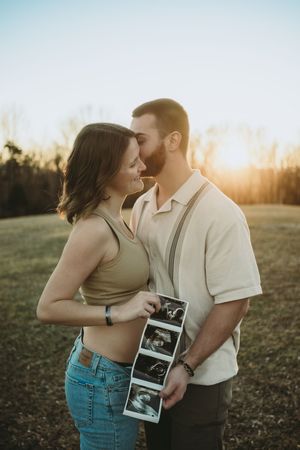 Couple holding ultrasound photos at sunset in a field, announcing pregnancy with glowing backlight.
