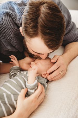 A loving father gently kisses his newborn baby dressed in a grey striped onesie while lying on a white surface.