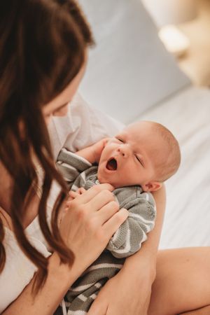 A mother tenderly holds her yawning newborn baby wrapped in a gray blanket while sitting on a bed.
