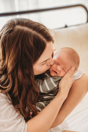 A mother tenderly nuzzles her newborn baby wrapped in a striped outfit while sitting on a bed.