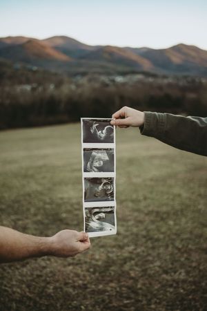 Two hands holding a strip of baby ultrasound photos with scenic mountain landscape in background.